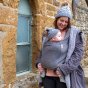 Woman stood in front of a stone wall holding her baby in the close caboo cotton blend baby carrier in the olive colour