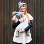 Woman stood in front of a metal wall holding a baby in the close caboo lite baby carrier in the blush cream colour 