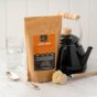 EcoLiving Citric Acid versatile household product. A natural food-grade cleaning product in brown plastic-free packaging. Recyclable and home compostable. Front lifestyle  view. White wooden background, kettle and brush in foreground. 