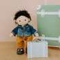 Olli ella dinkum doll stood on a wooden floor next to its sage travel tog case wearing a denim jacket and orange trousers