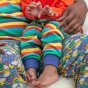close up of child wearing organic cotton baby and toddler bottoms with a bright rainbow stripe design with a co-ordinating blue waistband and stretchy cuffs that fold up from piccalilly