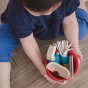 Child holding Plan Toys Cleaning Role Play Set including wooden squeegee, mop, dustpan cleaning spray in a red bucket. 