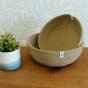 ReSpiin Jute Natural Medium & large woven Bowls with white respiin labels on a table with a tan sheet, a plant on the left and an off-white leafy wallpaper behind