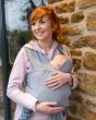 Woman leaning against a stone wall holding a baby in the close caboo lite baby carrier in the light grey alloy colour