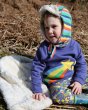 Young child sat on a fluffy blanket wearing the Piccalily rainbow sweatshirt and rainbow stripe flappy hat