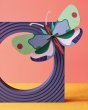 Studio roof slotting cardboard mint forest butterfly model climbing on a purple block sculpture in front of a pink wall