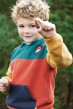 Child wearing the deep spruce colour block hoody in teal, rust red, mustard yellow and a navy pocket, holding a small pine cone to the camera with trees in the background