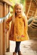 Smiling child wearing Frugi#s gold cord long sleeved button dress with floral appliques areound the hem, tights, and touching a fence