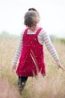 Child wearing Frugi's Greta berry dungaree dress with striped long sleeve top underneath, walking through a field