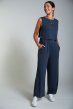 Smiling woman wearing the indigo Jessie maternity & nursing jumpsuit by Frugi with hands in pockets & white trainers on a grey background
