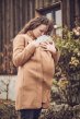 Mamalila Eco Wool Oslo Babywearing Coat in Camel. A light tan boiled wool winter babywearing coat. Front lifestyle view, caregiver carrying baby on the front. Wooden house background.  