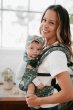 Woman wearing the Tula free to grow adjustable soft baby carrier, holding a small baby in a white room