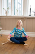 A child wearing the Maxomorra organic long sleeved top in Rainbow Farm with repeat rainbow and cloud pattern with a turquoise background and coordinated pink trim. Child is using a balance board, sitting cross legged