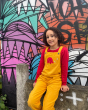 Girl sat on a wall in front of some graffiti wearing the Babipur x Frugi organic cotton pluto cord dungarees in the gold colour