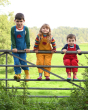 Three children stood on a metal gate outside, wearing the blue, yellow and red Frugi x Babipur Organic Cotton Cord Dungarees