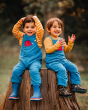 Two children happily sitting on a tree stump outside, both wearing the Blue Natural Organic Cord Dungarees by Frugi x Babipur, with a red elephant patch on the chest pocket