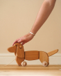 Close up of a hand reaching down to touch the Bajo natural wooden toy dachshund on a wooden floor