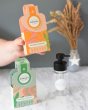 An adults hand holding up a pack of sanddorn scented Ben & Anna shampoo flakes,  the aloe vera scented shampoo flakes along with the dispenser bottle can be seen in the background 