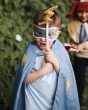 A close up of a child wearing the Avery Row Super Hero dress up set, showing the fabric lightening bolt wand and cape. Behind the child is another wearing a Jesters costume
