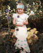 A child sitting on a rock outside, surrounded by beautiful wild flowers. The child is wearing the Avery Row Mermaid Dress Up Set that comprises of a light blue crown with shell details, and tail with blue scale details on the bottom. The child holds the w