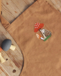 Avery Row Kid's Apron - Woodland Hedgehog. A beautiful light brown cotton canvas apron with a detailed, embroidered hedgehog and mushroom pattern of the chest of the apron, laid on a wooden table next to wooden, toy mushrooms.