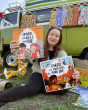 Person sat on some grass in front of a green van holding up the Omar, The Bees and Me and Enzo, The Swallows and Me children's books from Owlet Press