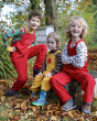 Three children happily sat outside on a wooden tree stump wearing the Red and Yellow Babipur x Frugi organic cotton cord dungarees. In the background are trees and a bee hive