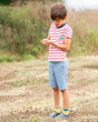 Young boy stood in a grass field wearing the Frugi reversible rainbow check rhys shorts