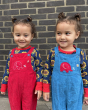 Two children smiling, and stood in front of a dark brick wall, wearing the red and yellow Frugi x Babipur organic cotton cord dungaress