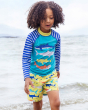 A child happily playing in the sea wearing the Frugi Boscastle Board Shorts - Banana Sharks, made from recycled bottles. A light banana yellow short with fun and colourful shark print, with a light blue draw string on the waist. This child is also wearing