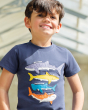 A child happily wearing the Frugi Avery Applique T-Shirt - Sharks. A deep Navy Blue short sleeve t-shirt made from GOTS organic cotton, with four colourful sharks sewn onto the front. From top to bottom is a lilac whale shark, a yellow great white shark, 