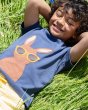 A child happily lying on the grass in the sun, wearing the Frugi Carsen Applique T-Shirt - Hare. A deep navy blue GOTS organic cotton t-shirt with a brown hare wearing yellow sunglasses with a rainbow reflection in the lens. The child is also wearing Yell