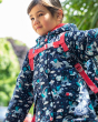 Young girl stood wearing the Frugi pegasus explorer rain jacket in front of a green tree