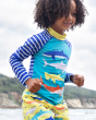 A happy child playing at the seaside, wearing the Frugi Rash Vest Sun Safe - Shark.  A blue sun-safe rash vest with a yellow neck, blue and white striped arms and fun shark print on the front, on a cream background. This rash vest passes British Sun-safe 