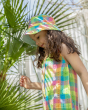 A child stood in front of a large green leafed plant, wearing the Frugi Organic Sorrel Reversible Hat - Summertime Check, which shows a little bit of the light blue underneath, and a Summertime Check outfit