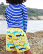 A child standing on large rocks at the beach wearing the Frugi Boscastle Board Shorts - Banana Sharks, made from recycled bottles. A light banana yellow short with fun and colourful shark print, with a light blue draw string on the waist. This child is al