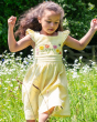 A child happily running in a flower field filled with white flowers, whilst wearing the Frugi Jasmine Dress - Flowers made from GOTS Organic cotton. A light sunshine, yellow and white striped short sleeve dress with adjustable ruffle straps, smocked back,