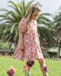 A child looking down at some flowers outside on a sunny day, wearing the Frugi GOTS organic cotton Matilda Collared Dress - Pink Floral Fun. A playful spring-time print of white and red flowers, bees and ladybirds on a light pink fabric. The dress is a Ti