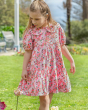 A child looking down at some flowers outside on a sunny day, wearing the Frugi GOTS organic cotton Matilda Collared Dress - Pink Floral Fun. A playful spring-time print of white and red flowers, bees and ladybirds on a light pink fabric. The dress is a Ti