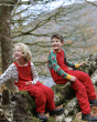 Two children happily sat outside in a tree, wearing the red Frugi x Babipur organic cotton cord dungarees