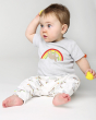 A happy toddler sat down and wearing the Frugi Children's GOTS Organic Cotton Frankie Summer Outfit - Sleepy Sloths. A beautiful 2 piece outfit including a grey short sleeved top with a sloth and rainbow character applique with comfy white cuffed sloth an