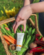 A person picking up the Green People Mineral Sun Cream SPF 15 with Natural Insect Repellent 100ml bottle from inside a basket full of colourful flowers and vegetables