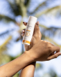A person holding the Green People Natural and Organic Mineral Suncream SPF30 Scent Free bottle in their hand, with a palm tree blurred in the background