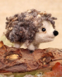The Makerss Needle Felt Small Curly Hedgehog. A beautifully crafted curly haired hedgehog with a cute cream face and legs, a black nose and curly spines stood on top of a fallen tree branch
