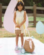 A child happily stood in the sun next to the Olli Ella Rattan Bunny Luggy with Lining – Gumdrop and holding the Rattan basket in Gumdrop