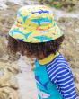 A child wearing the Frugi Harbour Swim Hat - Banana Sharks. A banana yellow sun hat with colourful shark print details, on a cream back ground. The hat is made from quick drying, recycled fabric that meets UPF 50+. The child also wears the Frugi Sun Safe 