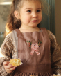A child eating a cake, whist wearing the Avery Row Kid's Pinafore Apron - Love Birds. A beautiful, dusky pink pinafore apron with two delicately embroidered pink love birds on the chest panel.