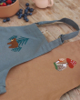 Avery Row Kid's Apron - Forest Bear. A beautiful blue cotton canvas apron with a detailed, embroidered bear and tree pattern of the chest of the apron, laid out on a wooden table with a bowl of fruit and a drink. Laid next to this apron is the light brown