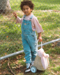 A child in blue dungarees and a pink top, stood next to a fence outside. The child is holding the handle of the Olli Ella Rattan Bunny Luggy with Lining – Pansy Floral. A beautifully woven pull along Luggy basket with cream cotton lining with a vintage pa