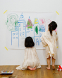 2 children crouched down drawing a town scene on a large piece of paper, taped to a wall